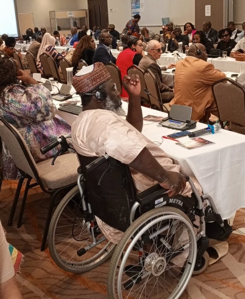 ADF Chair Idriss Maiga in a wheelchair participates in a conference session, surrounded by other attendees seated at tables covered with documents and laptops. Everyone appears engaged in the discussion.