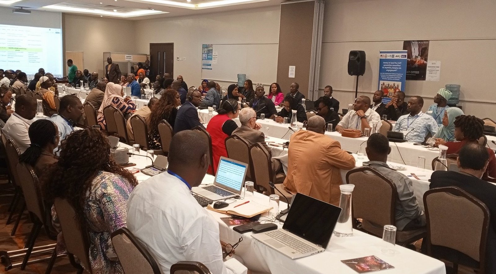 A large group of people sit in a conference room, attentively listening and taking notes. Some have laptops open. Posters and presentation slides are visible on the walls.