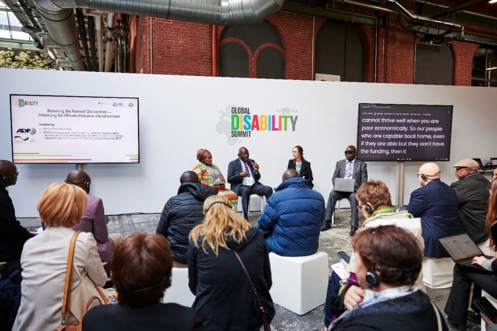 A panel discussion at the Global Disability Summit 2025 in Berlin features four speakers seated in front of a white backdrop with the summit’s colorful logo. The session, titled “Boosting the Nairobi Declaration – Alliancing for African Inclusive Development,” is hosted by the African Disability Forum (ADF) and partners, as shown on a screen to the left. Attendees, some wearing headsets for interpretation, are seated and listening intently. A second screen displays live captions of the speaker’s dialogue. The venue has an industrial design with exposed pipes and brick walls.