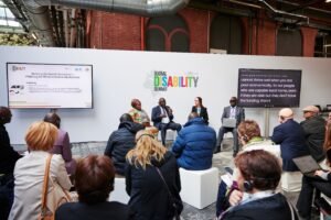 A panel discussion at the Global Disability Summit 2025 in Berlin features four speakers seated in front of a white backdrop with the summit’s colorful logo. The session, titled “Boosting the Nairobi Declaration – Alliancing for African Inclusive Development,” is hosted by the African Disability Forum (ADF) and partners, as shown on a screen to the left. Attendees, some wearing headsets for interpretation, are seated and listening intently. A second screen displays live captions of the speaker’s dialogue. The venue has an industrial design with exposed pipes and brick walls.