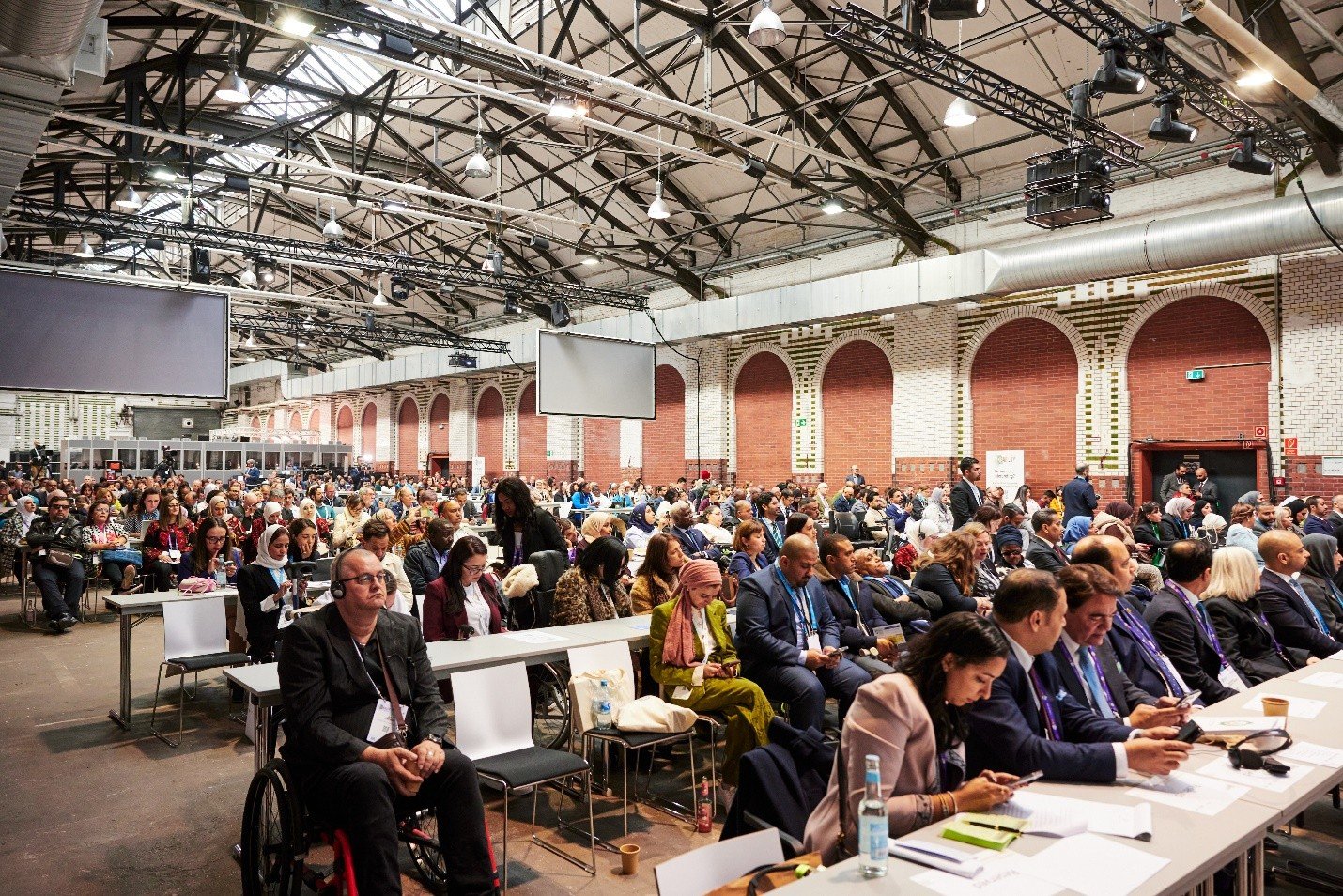 A large, diverse group of people attending a conference in a spacious industrial-style hall with high ceilings and exposed beams. Attendees are seated in rows, with some using wheelchairs. A panel of speakers sits at the front, and multiple large screens hang overhead. The atmosphere is professional and inclusive.