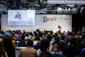 A speaker addresses a large audience at the 2025 Global Disability Summit in Berlin. The stage backdrop displays the summit’s logo along with partner logos, including the German Federal Government, IDA, and the Hashemite Kingdom of Jordan. A large screen projects the speaker's image with live captions and a sign language interpreter. The audience is seated and engaged, reflecting the event’s inclusive and international nature.