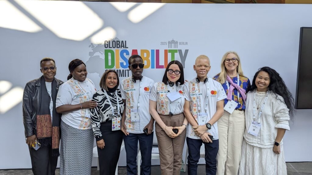 A diverse group of eight people, including youth delegates with disabilities and representatives from ADF, MasterCard Foundation, and Light for the World, pose in front of a banner for the Global Disability Summit 2025 in Berlin. Some wear white t-shirts with 'We Can Work' branding, while others wear professional or casual attire. A few participants have albinism, and some wear assistive devices like headsets or glasses. They are smiling and standing closely together.