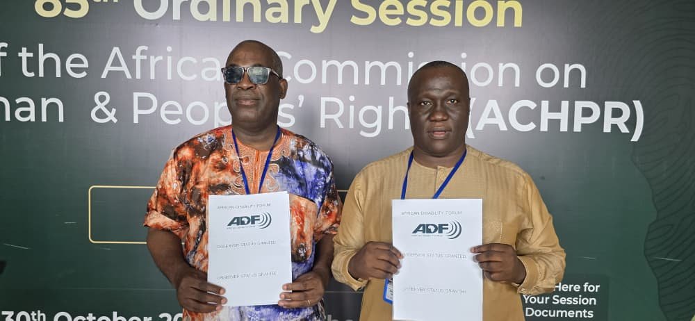Two men standing in front of a banner at the African Commission on Human & Peoples' Rights event, holding documents.