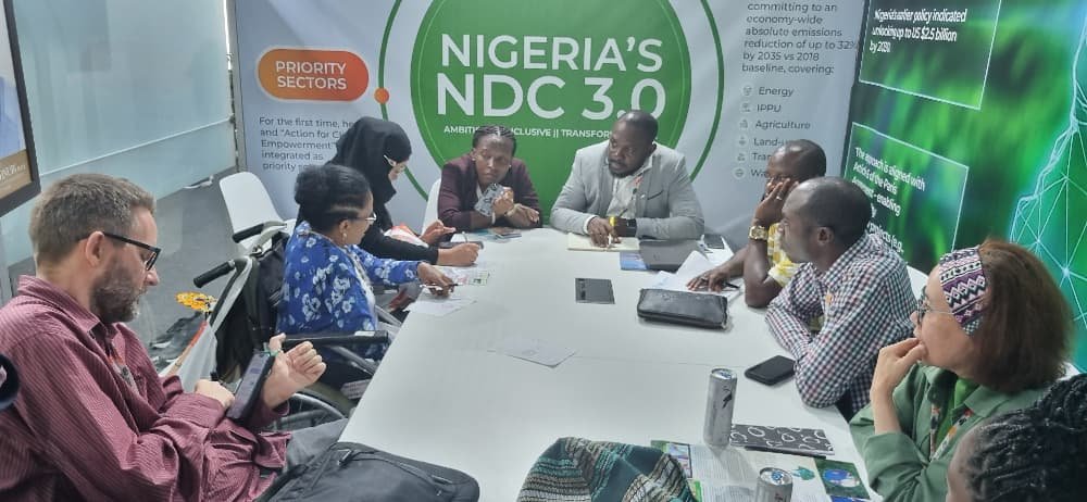 A diverse group of participants sit around a conference table engaged in a discussion. Several people are taking notes while others listen attentively. A green wall behind them displays text reading “Nigeria’s NDC 3.0.” The group includes persons with disabilities, and the setting appears to be a formal meeting room.