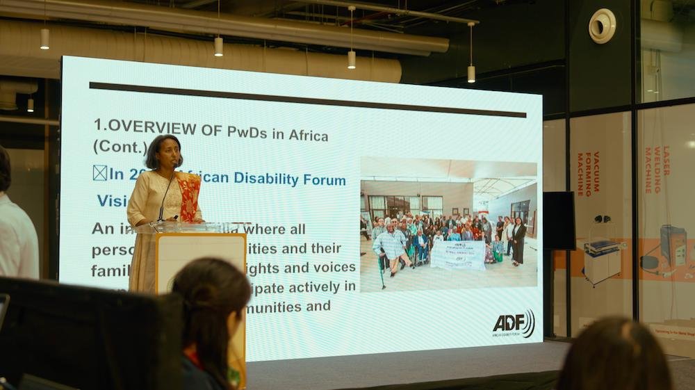 Bethelehem Zewde, ADF Programme Director, stands at a podium giving a presentation. Behind her, a large screen displays a slide titled “Overview of PwDs in Africa,” with text about the ADF vision and a group photo. Audience members are seated in the foreground, listening attentively in a modern conference room.