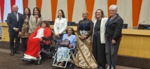 A diverse group of nine people, including two wheelchair users, smiling and posing together in a conference room with vibrant orange and beige paneling.