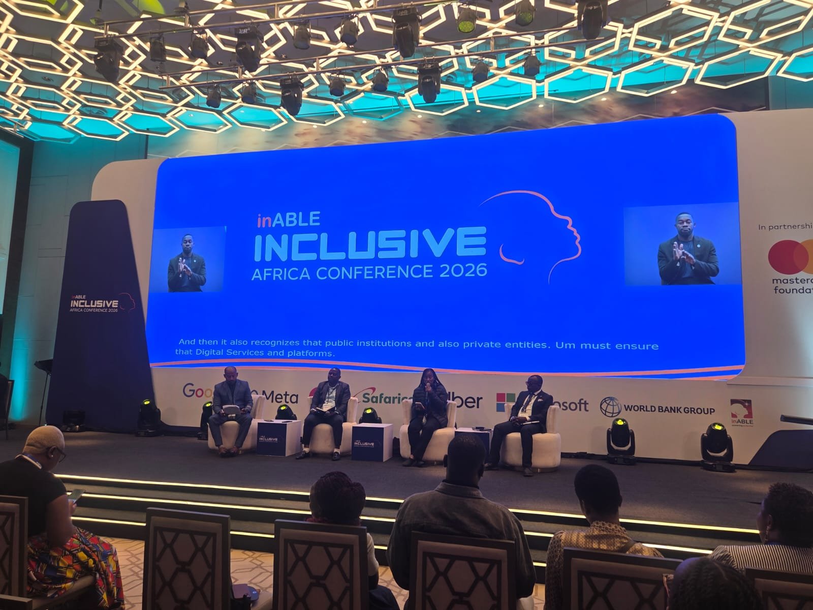 A conference stage with four speakers seated under a large blue screen displaying "inABLE Inclusive Africa Conference 2026." Audience members are in the foreground.