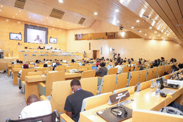 A spacious modern conference hall filled with attendees seated at wooden desks equipped with microphones and nameplates. A large screen at the front shows a speaker, with two smaller screens on either side. The ceiling is curved with recessed lighting. Country nameplates such as “Kenya,” “Lesotho,” and “Malawi” are visible, suggesting an international or regional meeting.