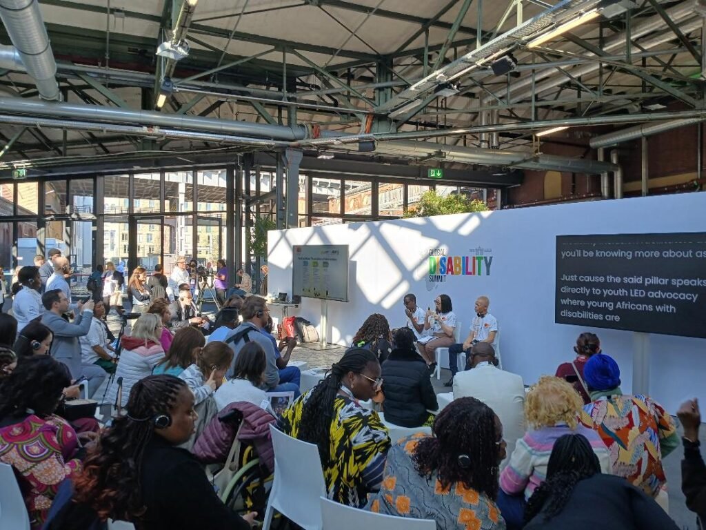A diverse audience, including people with disabilities, attentively listens to a fireside chat on economic inclusion at the Global Disability Summit 2025 in Berlin. The panelists, seated on a small stage against a backdrop with the summit’s logo, include young disability advocates wearing 'We Can Work' t-shirts. Many attendees wear headsets for translation or accessibility, and some are using wheelchairs. A large screen displays live captions of the discussion. The venue is an industrial-style space with high ceilings, large windows, and natural light streaming in.