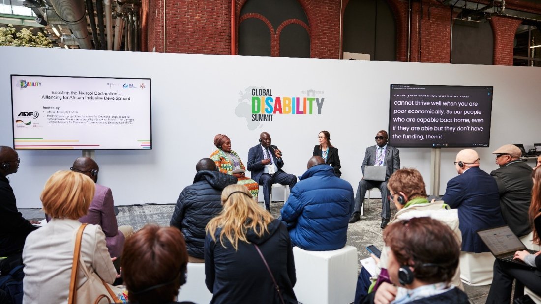 A panel discussion at the Global Disability Summit 2025 in Berlin features four speakers seated in front of a white backdrop with the summit’s colorful logo. The session, titled “Boosting the Nairobi Declaration – Alliancing for African Inclusive Development,” is hosted by the African Disability Forum (ADF) and partners, as shown on a screen to the left. Attendees, some wearing headsets for interpretation, are seated and listening intently. A second screen displays live captions of the speaker’s dialogue. The venue has an industrial design with exposed pipes and brick walls.