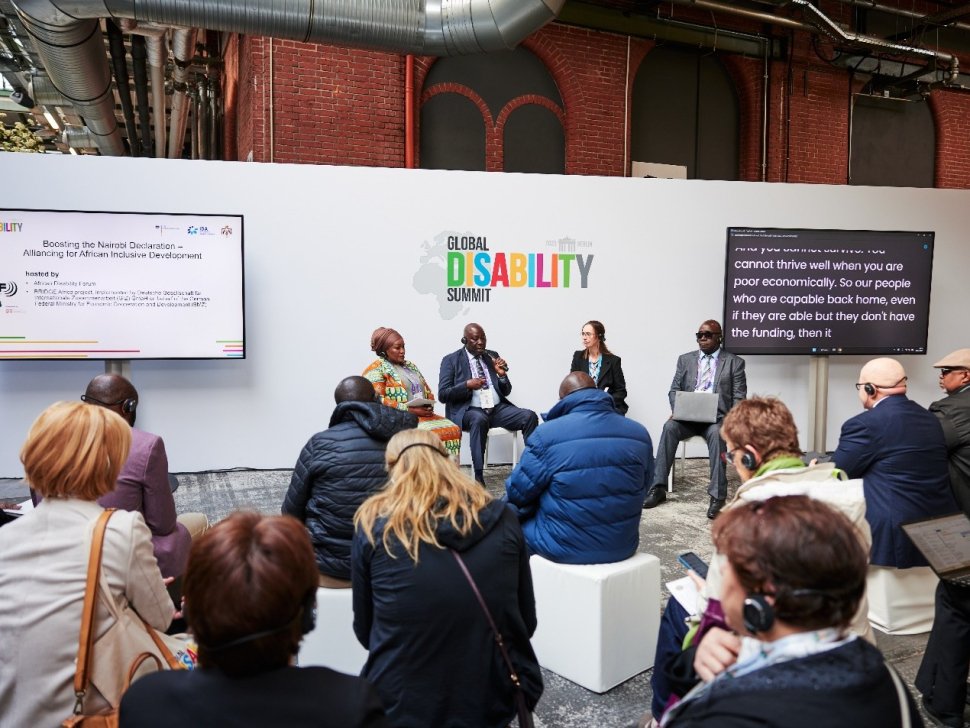 A panel discussion at the Global Disability Summit 2025 in Berlin features four speakers seated in front of a white backdrop with the summit’s colorful logo. The session, titled “Boosting the Nairobi Declaration – Alliancing for African Inclusive Development,” is hosted by the African Disability Forum (ADF) and partners, as shown on a screen to the left. Attendees, some wearing headsets for interpretation, are seated and listening intently. A second screen displays live captions of the speaker’s dialogue. The venue has an industrial design with exposed pipes and brick walls.