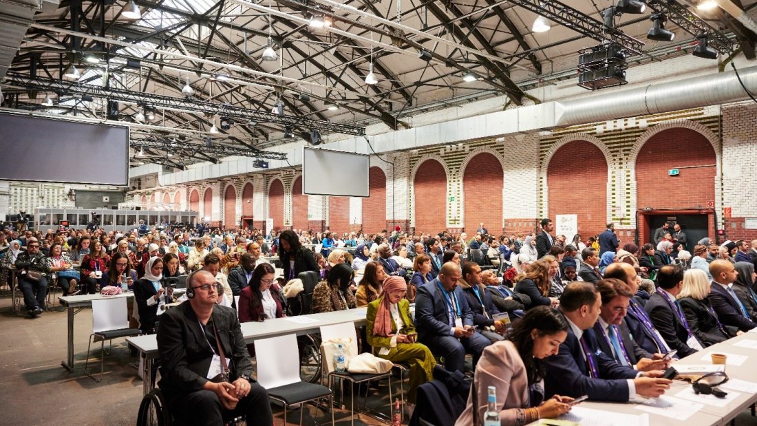 A large, diverse group of people attending a conference in a spacious industrial-style hall with high ceilings and exposed beams. Attendees are seated in rows, with some using wheelchairs. A panel of speakers sits at the front, and multiple large screens hang overhead. The atmosphere is professional and inclusive.