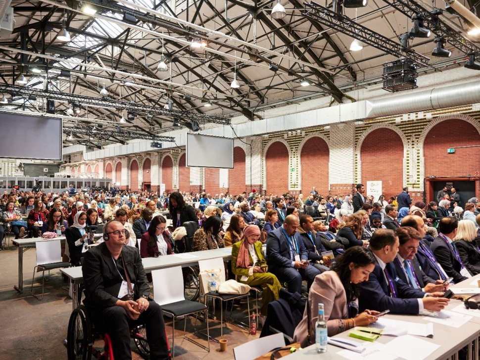 A large, diverse group of people attending a conference in a spacious industrial-style hall with high ceilings and exposed beams. Attendees are seated in rows, with some using wheelchairs. A panel of speakers sits at the front, and multiple large screens hang overhead. The atmosphere is professional and inclusive.