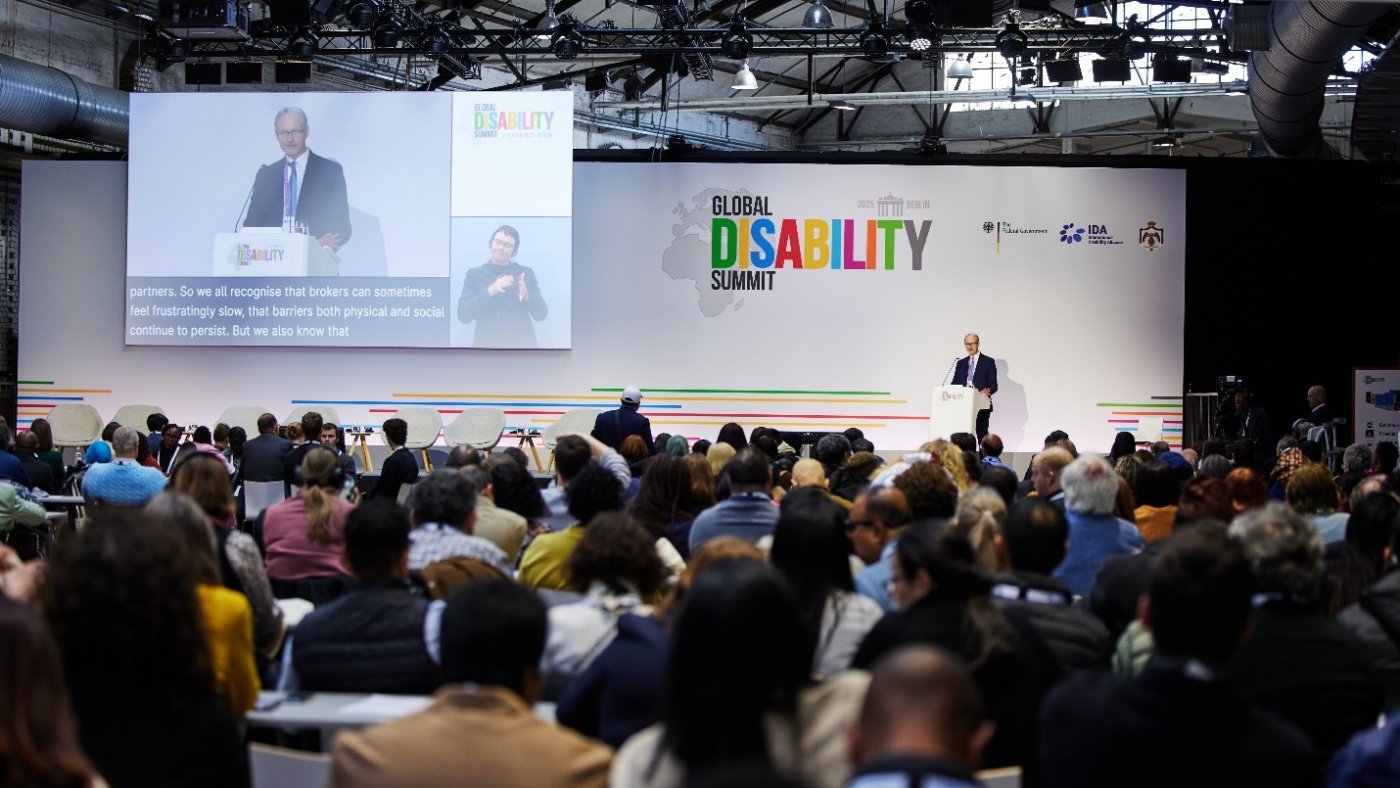 A speaker addresses a large audience at the 2025 Global Disability Summit in Berlin. The stage backdrop displays the summit’s logo along with partner logos, including the German Federal Government, IDA, and the Hashemite Kingdom of Jordan. A large screen projects the speaker's image with live captions and a sign language interpreter. The audience is seated and engaged, reflecting the event’s inclusive and international nature.