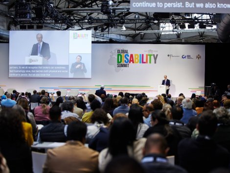 A speaker addresses a large audience at the 2025 Global Disability Summit in Berlin. The stage backdrop displays the summit’s logo along with partner logos, including the German Federal Government, IDA, and the Hashemite Kingdom of Jordan. A large screen projects the speaker's image with live captions and a sign language interpreter. The audience is seated and engaged, reflecting the event’s inclusive and international nature.