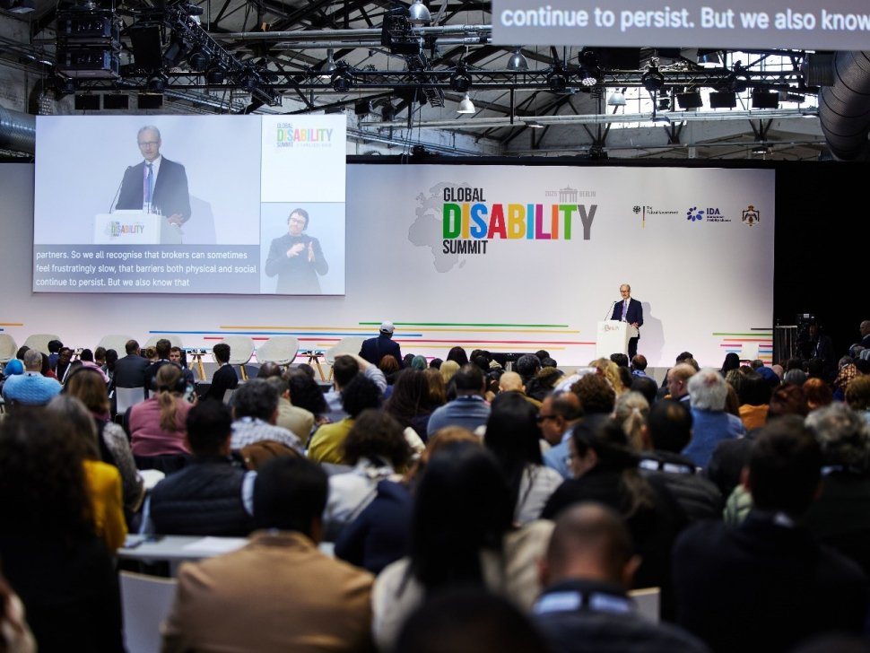 A speaker addresses a large audience at the 2025 Global Disability Summit in Berlin. The stage backdrop displays the summit’s logo along with partner logos, including the German Federal Government, IDA, and the Hashemite Kingdom of Jordan. A large screen projects the speaker's image with live captions and a sign language interpreter. The audience is seated and engaged, reflecting the event’s inclusive and international nature.