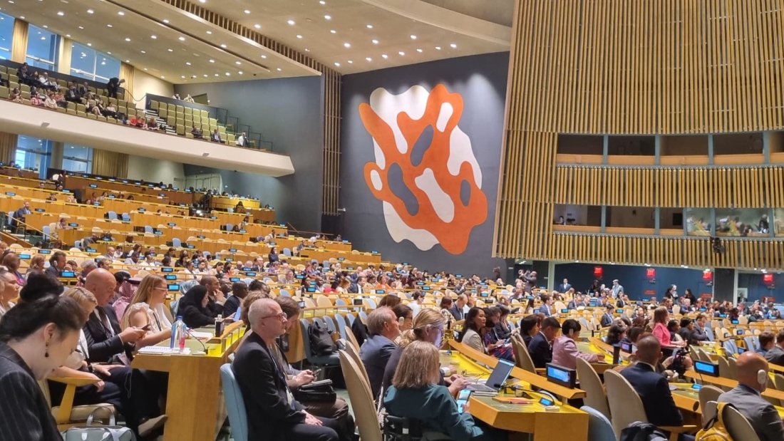 A wide view of the packed UN General Assembly Hall during COSP18, with delegates from around the world seated at rows of desks. The iconic orange and white mural is visible in the background, and several attendees, including wheelchair users, are engaged in the proceedings.|Sweden Sverige logo|light for the world logo|disabled people organization Denmark logo|humanity inclusion logo