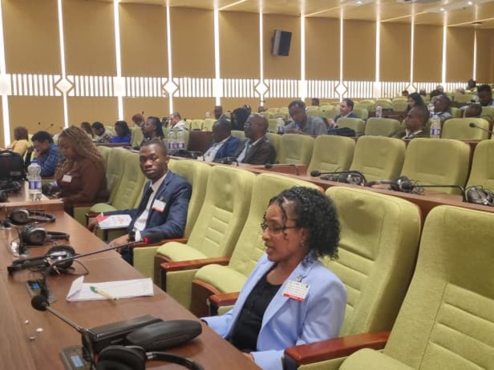 Shitaye Astawes, wearing a light blue jacket, speaks into a microphone while seated in a large conference hall with other participants in the background.