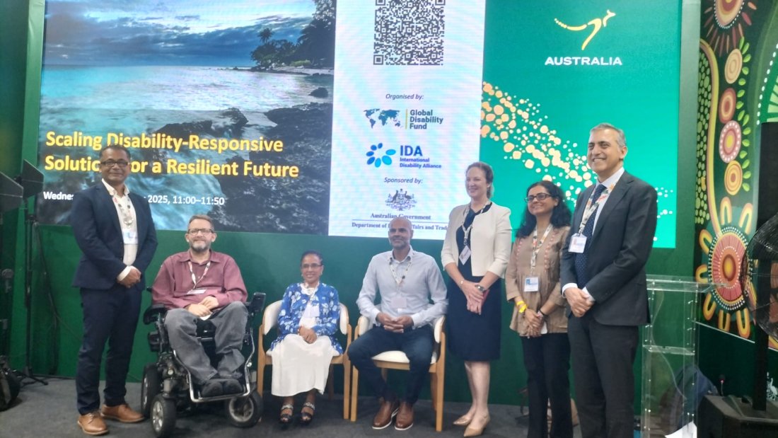 group of eight panelists and speakers pose on a stage in front of a large screen reading “Scaling Disability-Responsive Solutions for a Resilient Future.” The backdrop includes logos of the Global Disability Fund, the International Disability Alliance, and the Australian Government. Some participants are seated, including wheelchair users, while others stand, all wearing conference badges.|||
