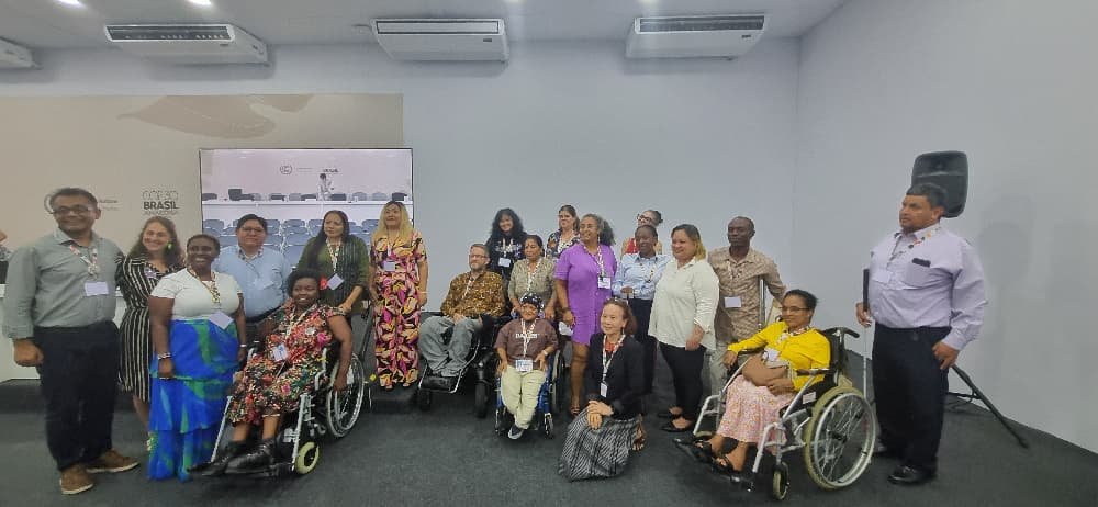 A large group of conference participants pose together in a meeting room for a group photo. The group includes women and men of diverse backgrounds, with several wheelchair users seated in front. Most participants are wearing name badges, and the room has a conference setup with chairs and a presentation screen in the background.