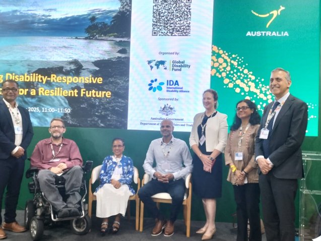 group of eight panelists and speakers pose on a stage in front of a large screen reading “Scaling Disability-Responsive Solutions for a Resilient Future.” The backdrop includes logos of the Global Disability Fund, the International Disability Alliance, and the Australian Government. Some participants are seated, including wheelchair users, while others stand, all wearing conference badges.|||
