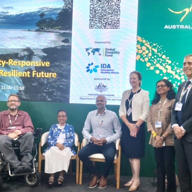 group of eight panelists and speakers pose on a stage in front of a large screen reading “Scaling Disability-Responsive Solutions for a Resilient Future.” The backdrop includes logos of the Global Disability Fund, the International Disability Alliance, and the Australian Government. Some participants are seated, including wheelchair users, while others stand, all wearing conference badges.|||
