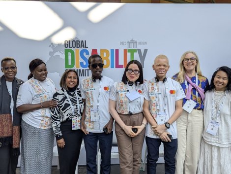 A diverse group of eight people, including youth delegates with disabilities and representatives from ADF, MasterCard Foundation, and Light for the World, pose in front of a banner for the Global Disability Summit 2025 in Berlin. Some wear white t-shirts with 'We Can Work' branding, while others wear professional or casual attire. A few participants have albinism, and some wear assistive devices like headsets or glasses. They are smiling and standing closely together.|