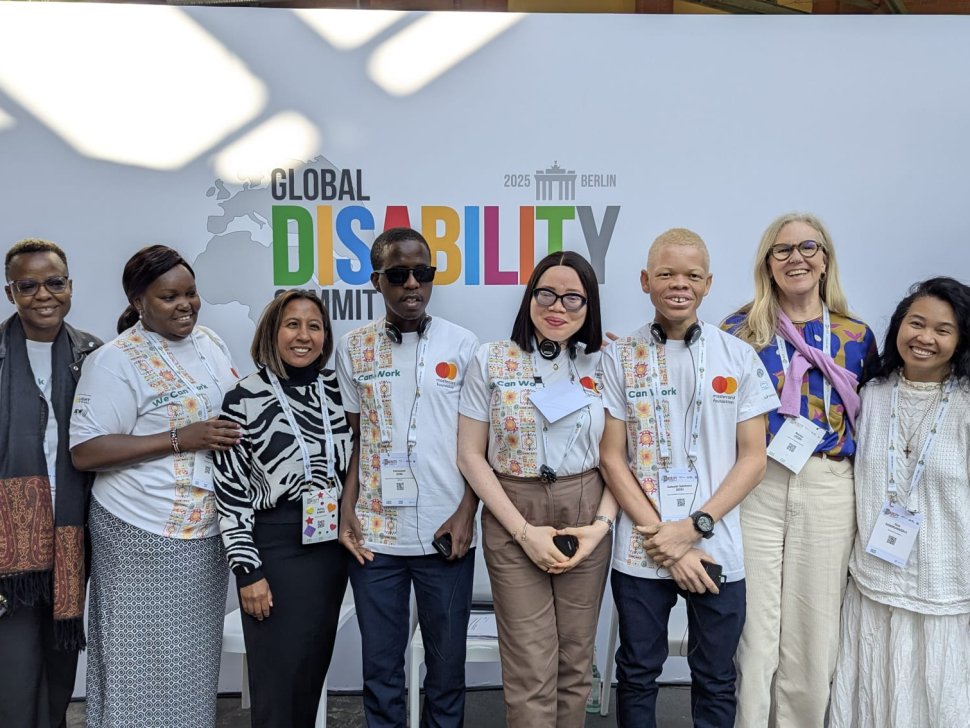 A diverse group of eight people, including youth delegates with disabilities and representatives from ADF, MasterCard Foundation, and Light for the World, pose in front of a banner for the Global Disability Summit 2025 in Berlin. Some wear white t-shirts with 'We Can Work' branding, while others wear professional or casual attire. A few participants have albinism, and some wear assistive devices like headsets or glasses. They are smiling and standing closely together.|